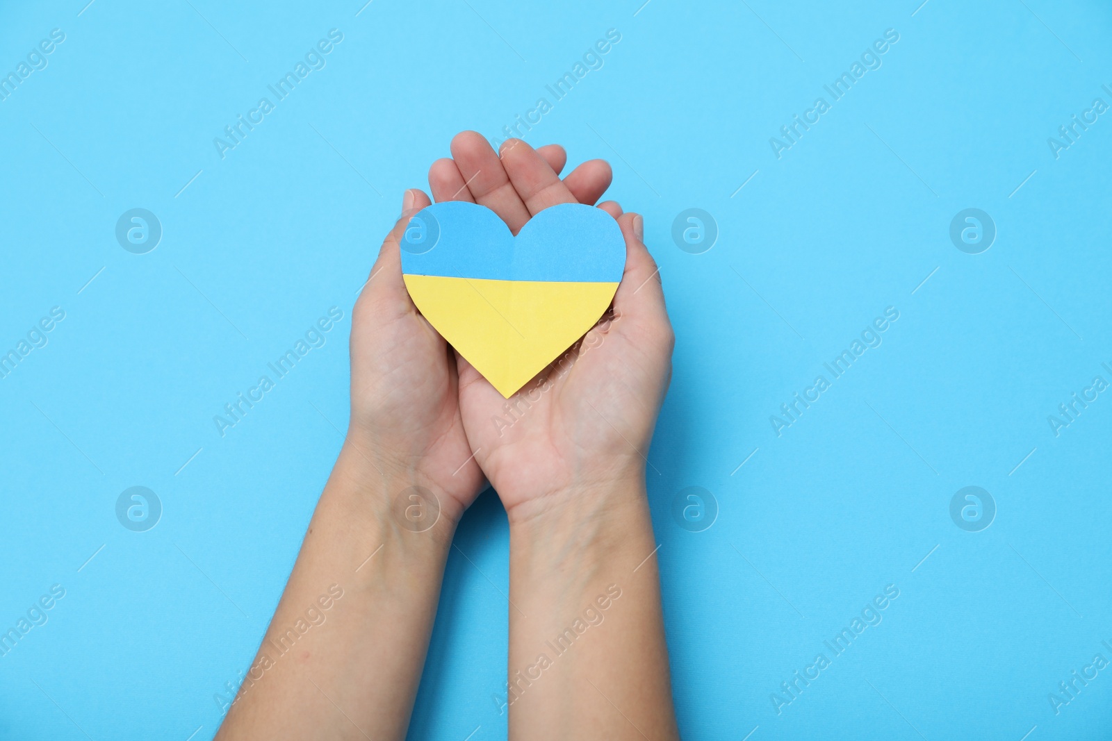 Photo of Woman holding paper heart in colors of Ukrainian flag on light blue background, top view