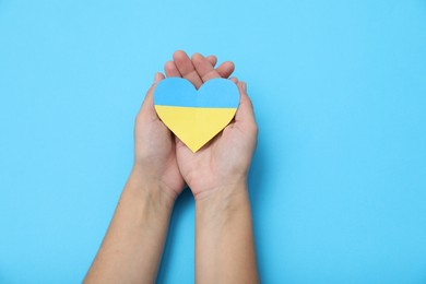 Photo of Woman holding paper heart in colors of Ukrainian flag on light blue background, top view