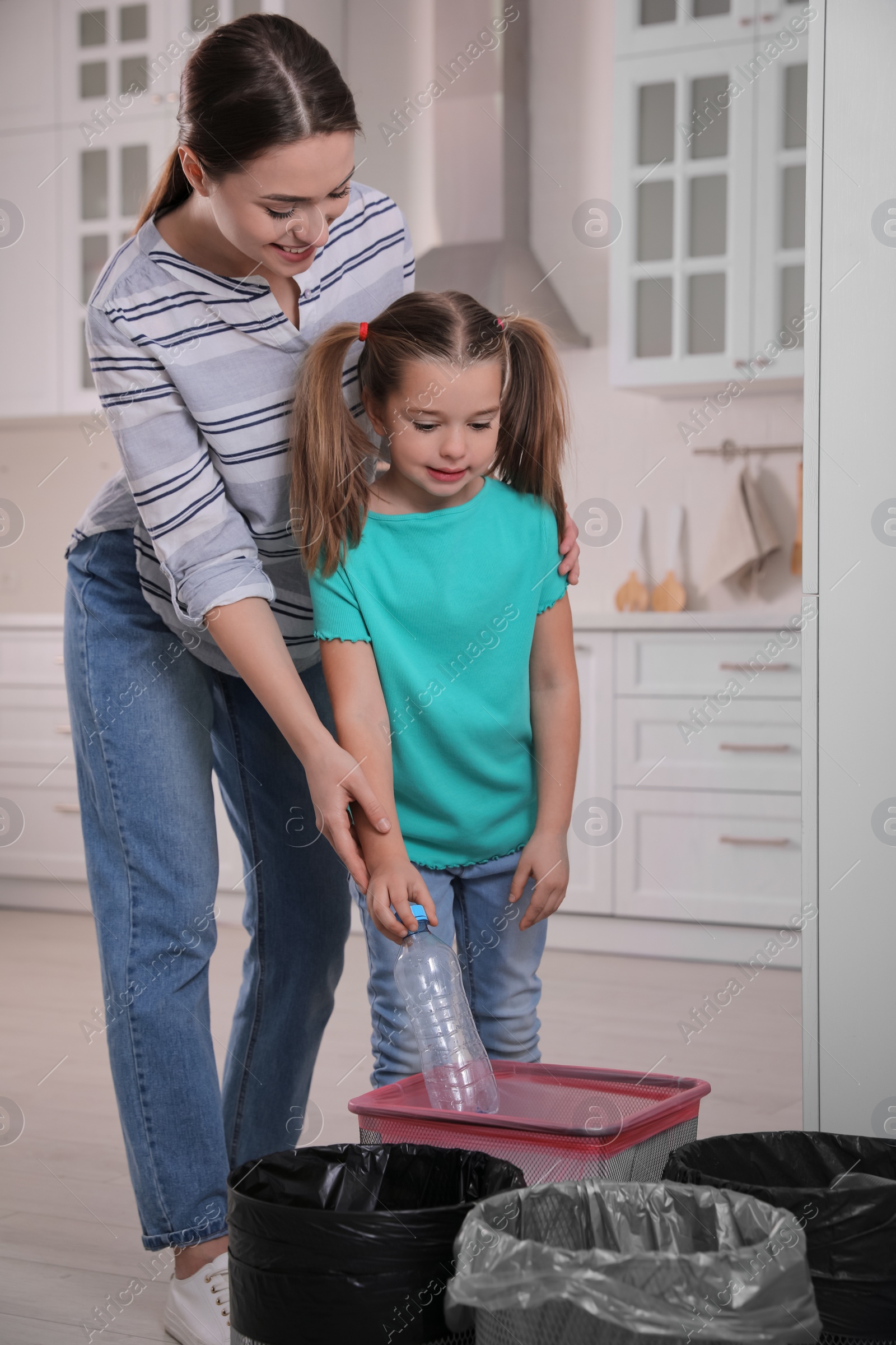 Photo of Young woman and her daughter throwing plastic bottle into trash bin in kitchen. Separate waste collection