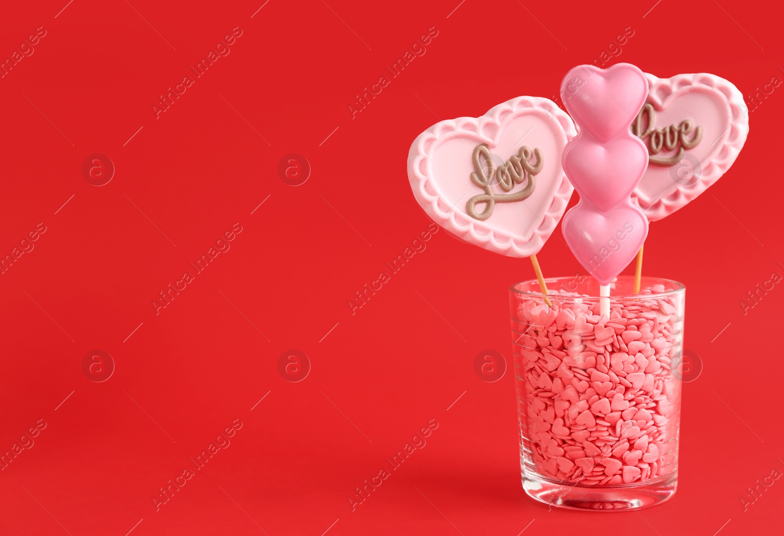 Photo of Different chocolate heart shaped lollipops in glass with sprinkles on red background. Space for text