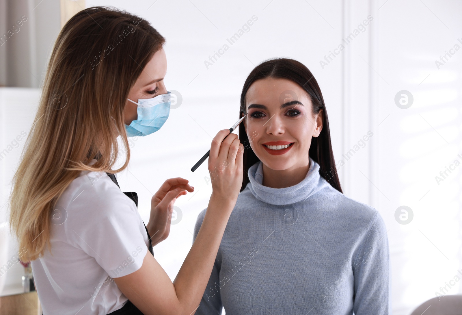 Photo of Makeup artist in protective mask working with woman indoors. Preventive measures during COVID-19 pandemic