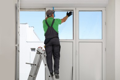 Worker on folding ladder installing window indoors, back view