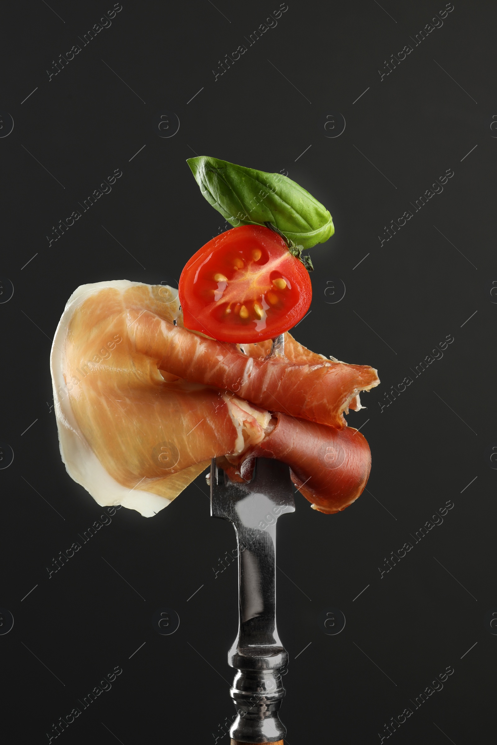 Photo of Fork with slice of tasty jamon, basil and tomato on black background, closeup