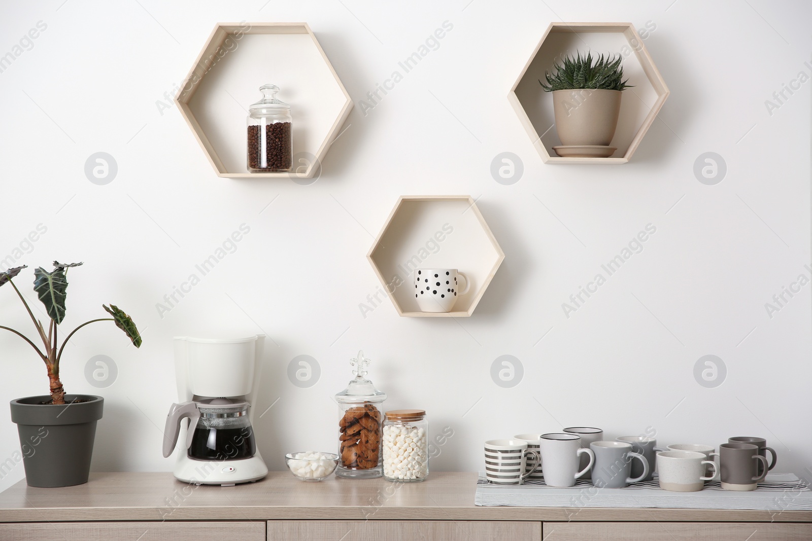 Photo of Modern coffeemaker and cups on commode near light wall indoors