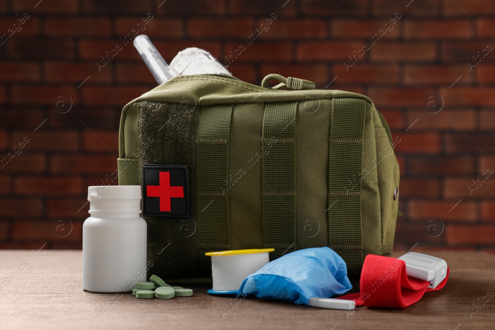 Photo of Military first aid kit, tourniquet, pills and elastic bandage on wooden table against brick wall