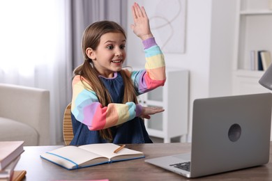 Photo of E-learning. Emotional girl raising her hand to answer during online lesson at table indoors