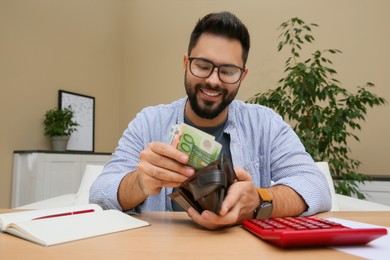 Photo of Young man putting money into wallet at table indoors