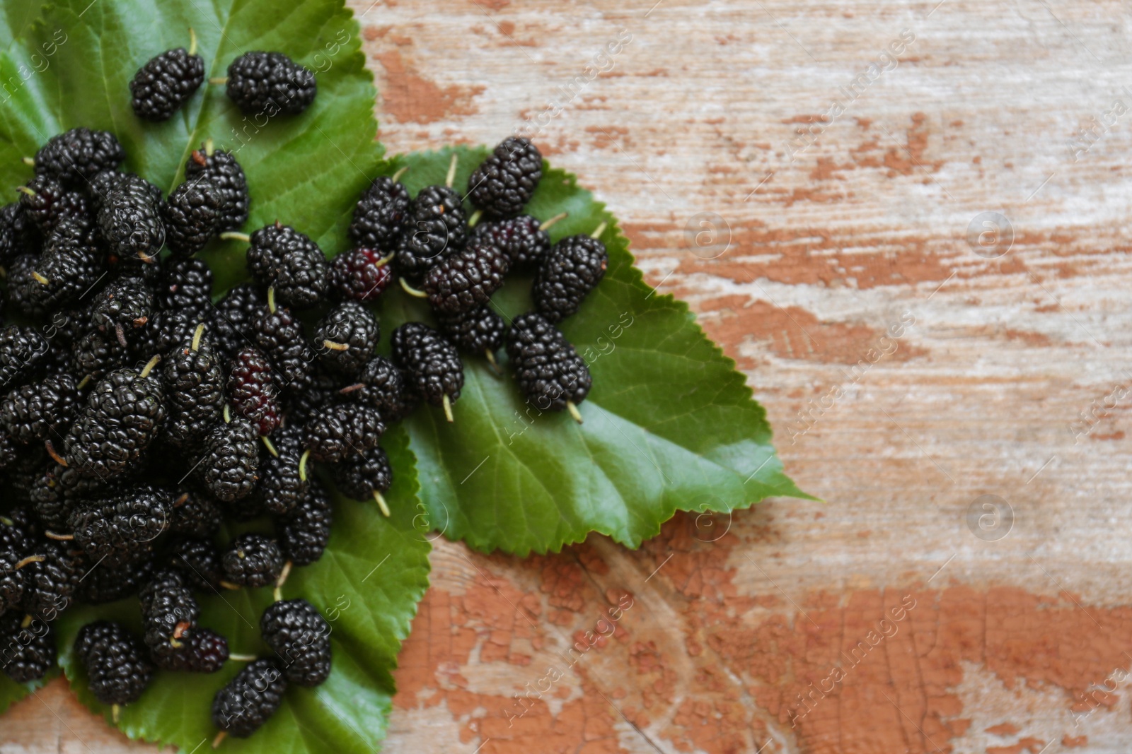 Photo of Heap of delicious ripe black mulberries and green leaves on wooden table, flat lay. Space for text