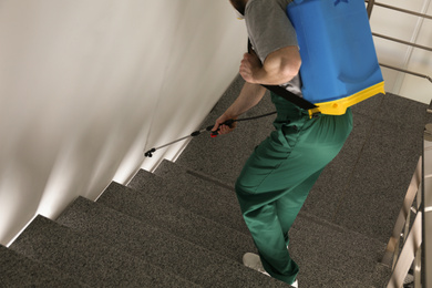 Photo of Pest control worker in uniform spraying pesticide on stairs indoors