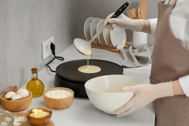 Woman cooking delicious crepe on electric pancake maker at white marble table in kitchen, closeup