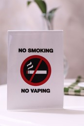 Photo of No Smoking No Vaping sign on white table indoors, closeup