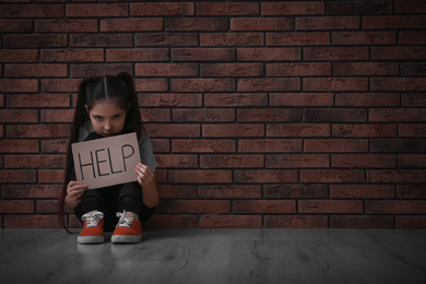 Sad little girl with sign HELP on floor near brick wall, space for text. Child in danger
