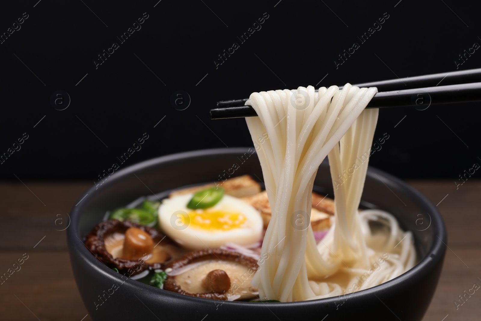 Photo of Eating delicious vegetarian ramen with chopsticks at table against black background, closeup