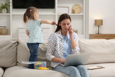 Photo of Woman working remotely at home. Little daughter bothering her mother on sofa in living room