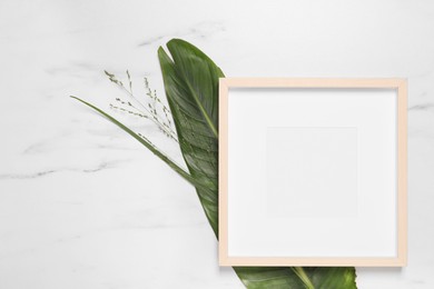 Photo of Empty photo frame, green leaf and flowers on white marble background, flat lay. Space for design