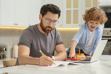 Photo of Father working remotely while his son playing with toys at home. Man writing in notebook at desk