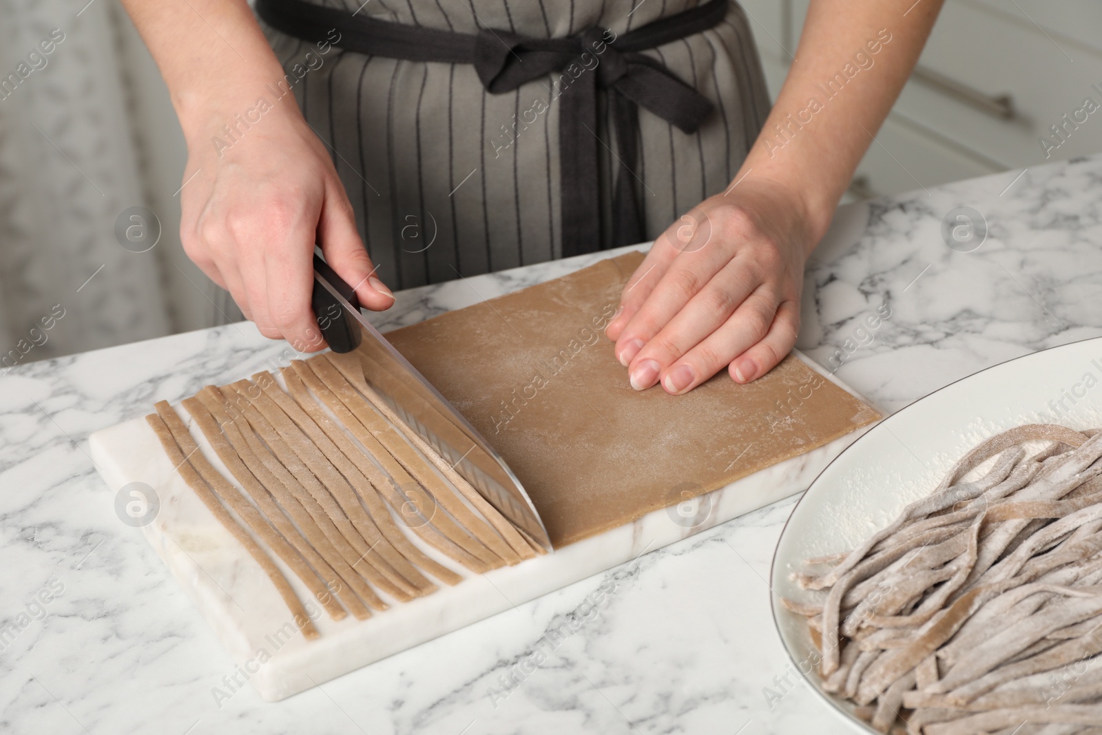 Photo of Woman making soba (buckwheat noodles) with knife at white marble table indoors, closeup