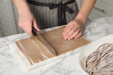 Photo of Woman making soba (buckwheat noodles) with knife at white marble table indoors, closeup