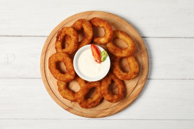 Wooden board with tasty onion rings and sauce on table, top view