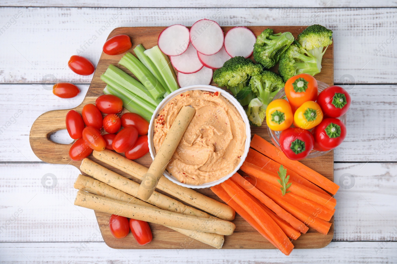 Photo of Board with delicious hummus, grissini sticks and fresh vegetables on white wooden background, top view