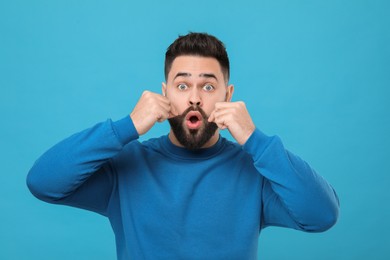 Surprised young man touching mustache on light blue background