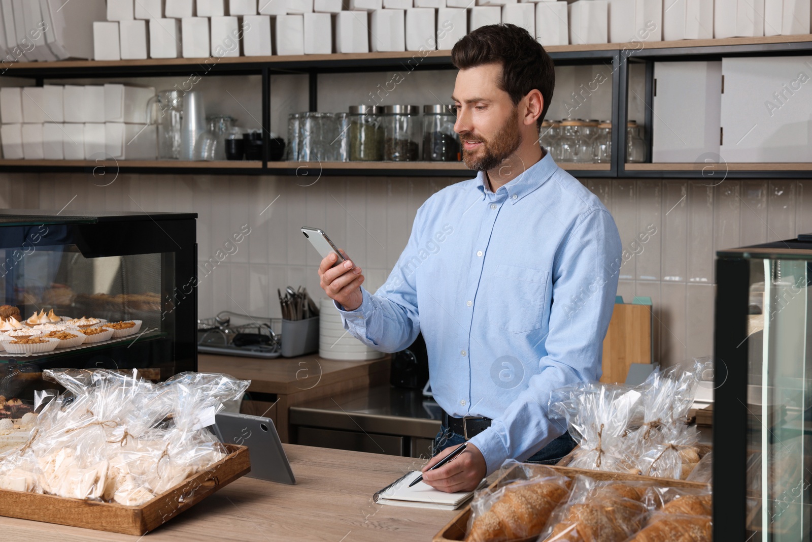 Photo of Handsome business owner with smartphone and pen at cashier desk in bakery shop