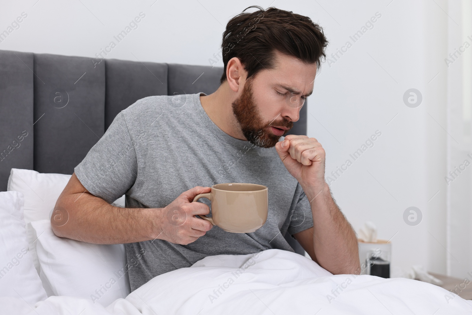 Photo of Sick man with cup of hot drink coughing on bed at home. Cold symptoms