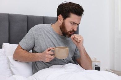 Sick man with cup of hot drink coughing on bed at home. Cold symptoms