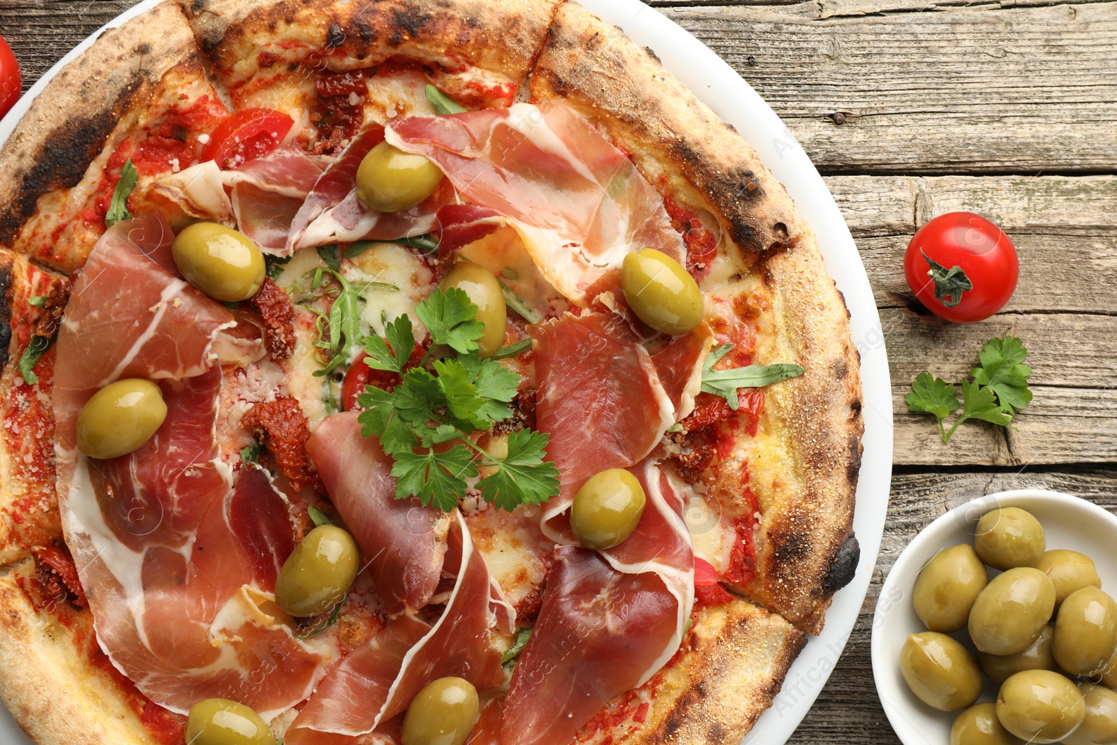 Photo of Tasty pizza with cured ham, olives, tomato and parsley on wooden table, top view