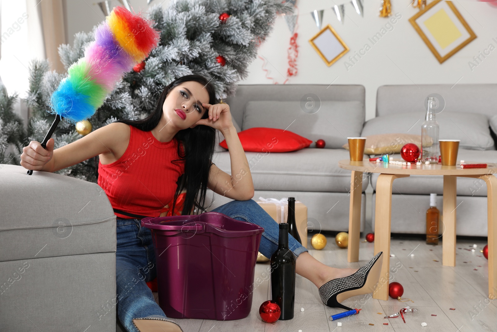 Photo of Young woman with dusting brush suffering from hangover in messy room after New Year party