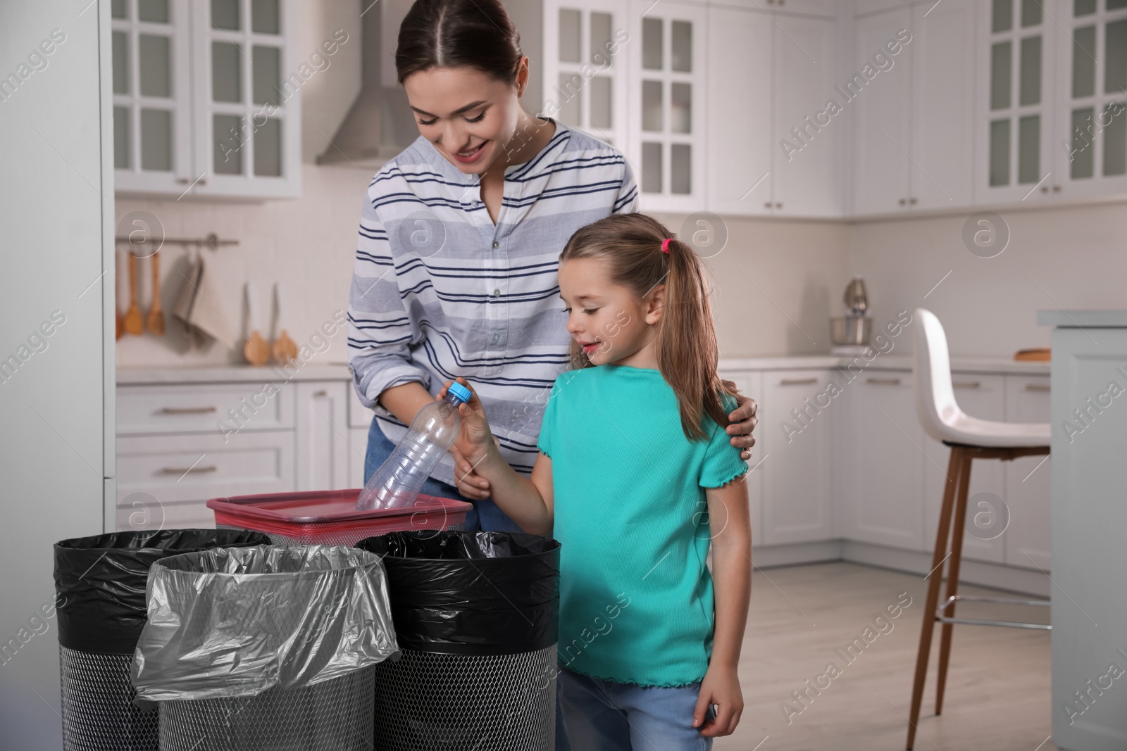 Photo of Young woman and her daughter throwing plastic bottle into trash bin in kitchen. Separate waste collection