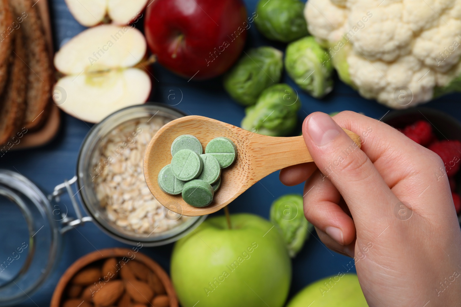 Photo of Woman holding wooden spoon of pills at table with foodstuff, top view. Prebiotic supplements