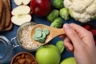 Photo of Woman holding wooden spoon of pills at table with foodstuff, top view. Prebiotic supplements