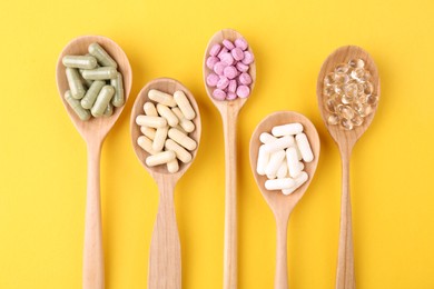 Different vitamin pills in wooden spoons on yellow background, flat lay