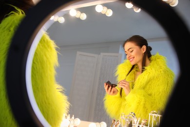 Young woman applying make up near illuminated mirror indoors, view through ring lamp