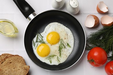 Frying pan with tasty cooked eggs, dill and other products on white tiled table, flat lay