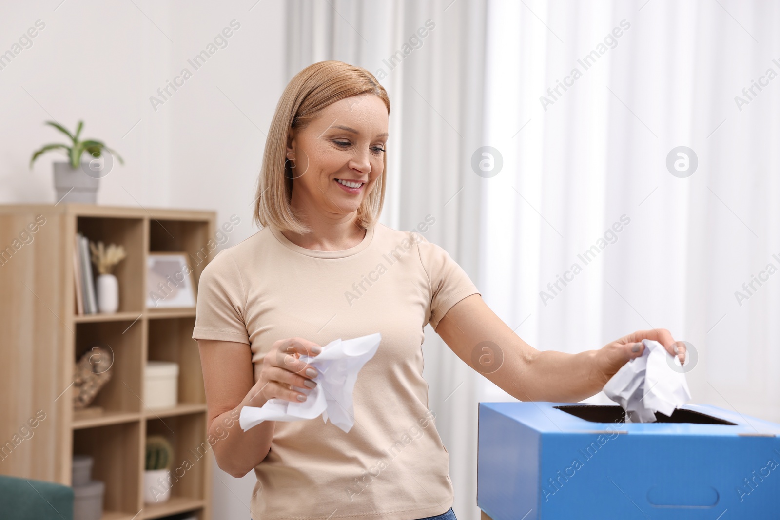 Photo of Garbage sorting. Smiling woman throwing crumpled paper into cardboard box in room