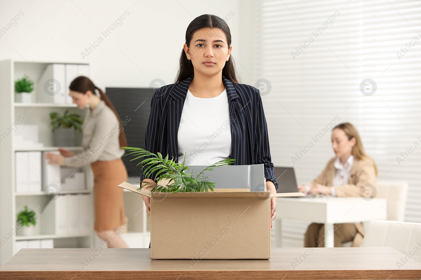 Photo of Unemployment problem. Frustrated woman with box of personal belongings at table in office