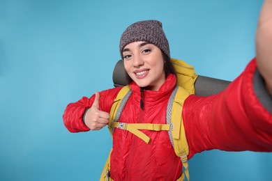 Photo of Smiling young woman with backpack taking selfie and showing thumb up on light blue background. Active tourism