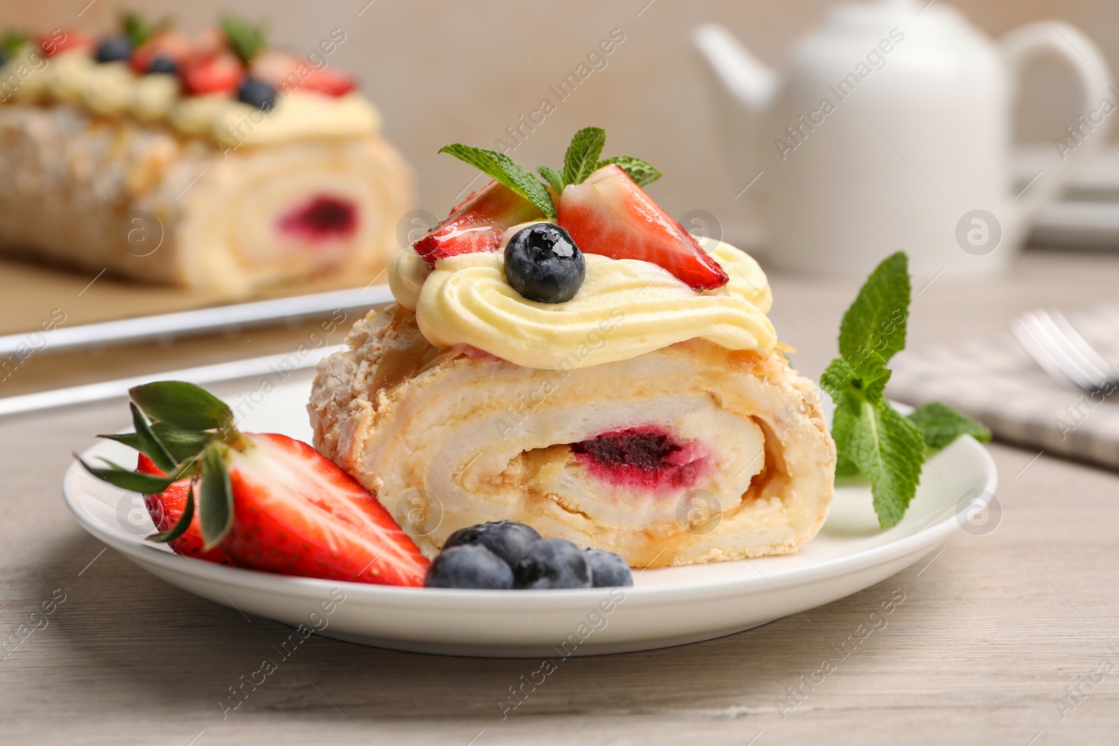 Photo of Piece of tasty meringue roll with jam, cream, strawberry, blueberry and mint on white wooden table, closeup