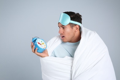 Emotional man in sleeping mask wrapped with blanket holding alarm clock on grey background