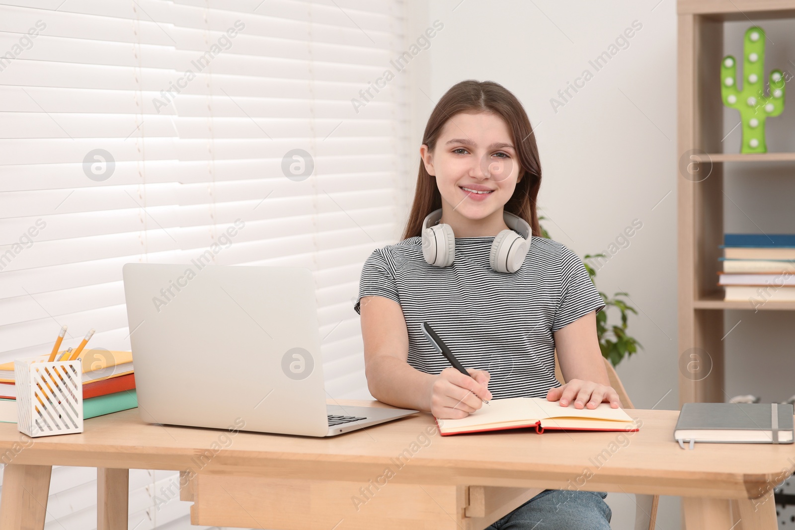 Photo of Cute girl with headphones writing in notepad near laptop at desk in room. Home workplace