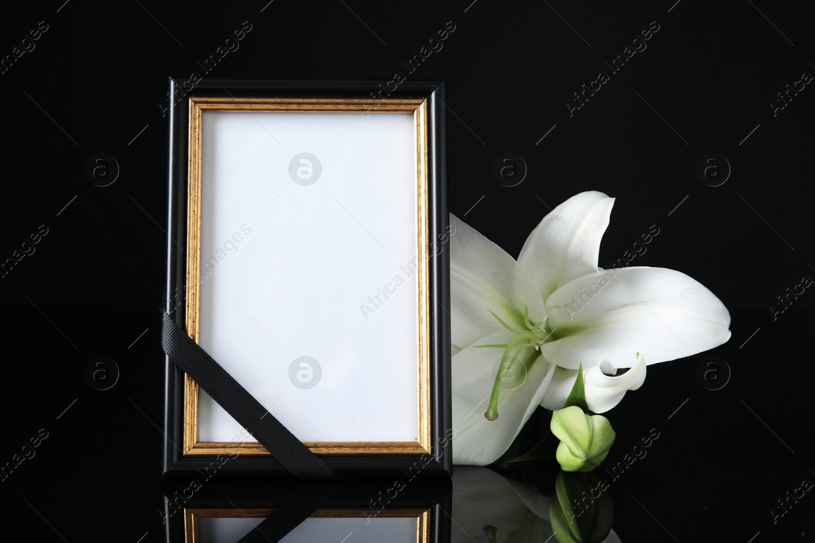 Photo of Funeral photo frame with ribbon and white lily on black table against dark background. Space for design