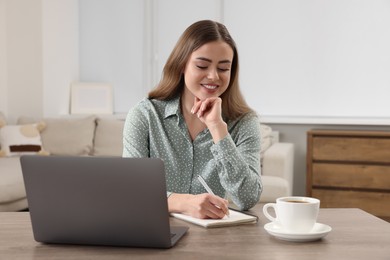 Happy woman writing something in notebook near laptop at wooden table in room