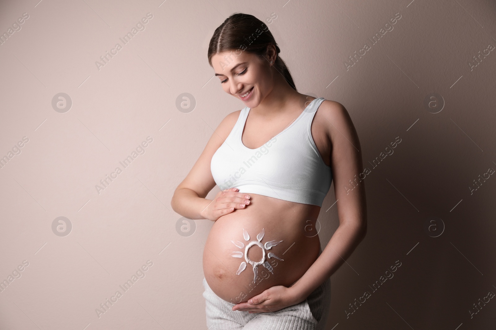 Photo of Young pregnant woman with sun protection cream on belly near beige wall