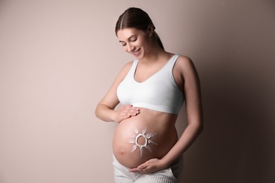 Photo of Young pregnant woman with sun protection cream on belly near beige wall