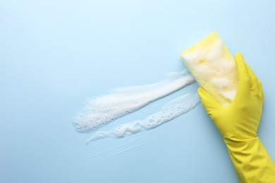 Photo of Cleaner in rubber glove holding sponge with foam on light blue background, top view