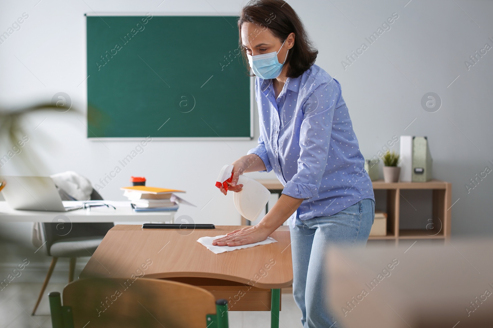 Photo of Teacher with protective mask disinfecting desk in classroom. Reopening after Covid-19 quarantine