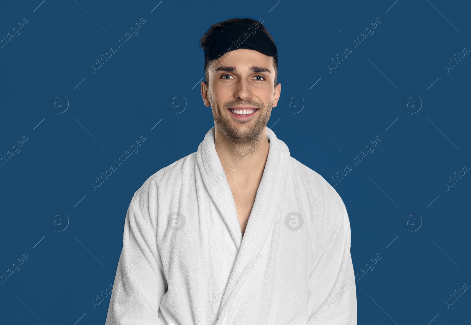 Photo of Happy young man in bathrobe and eye sleeping mask on blue background