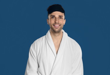 Happy young man in bathrobe and eye sleeping mask on blue background